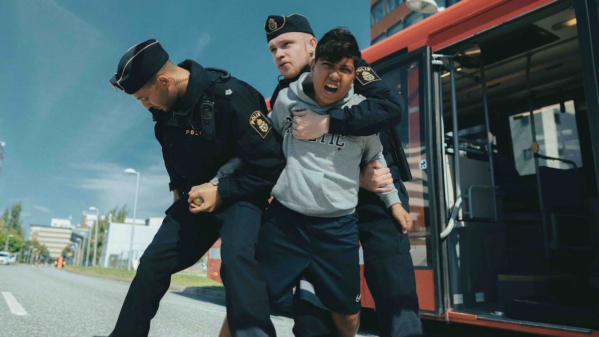 Syntolkning: En ung pojke brottas med två poliser utanför en röd buss.