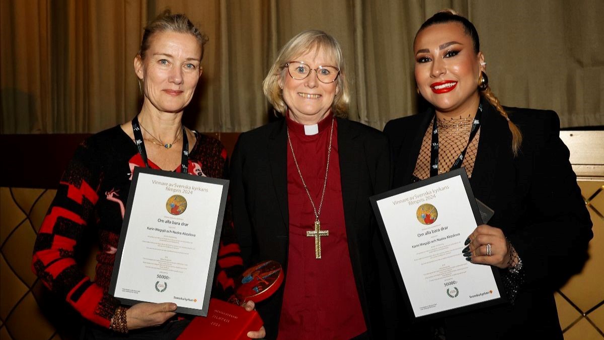 Syntolkning: Tre kvinnor ler mot kameran, en i biskopskläder och två med varsitt diplom.