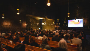 Syntolkning: Fulla kyrkbänkar i förgrunden, filmduk med texten "Tempo Dokumentärfestival" i bakgrunden.