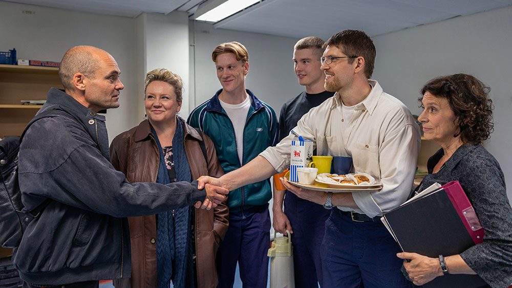 Syntolkning: David Dencik som Lars Norén skakar hand med internerna i ett kontorsrum.