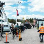 Filmkamera på ett högt stativ med Strandvägen i Stockholm i bakgrunden.