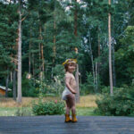 En bebis i blöja och med en blomsterkrans på huvudet står på en veranda med tallskog i bakgrunden.