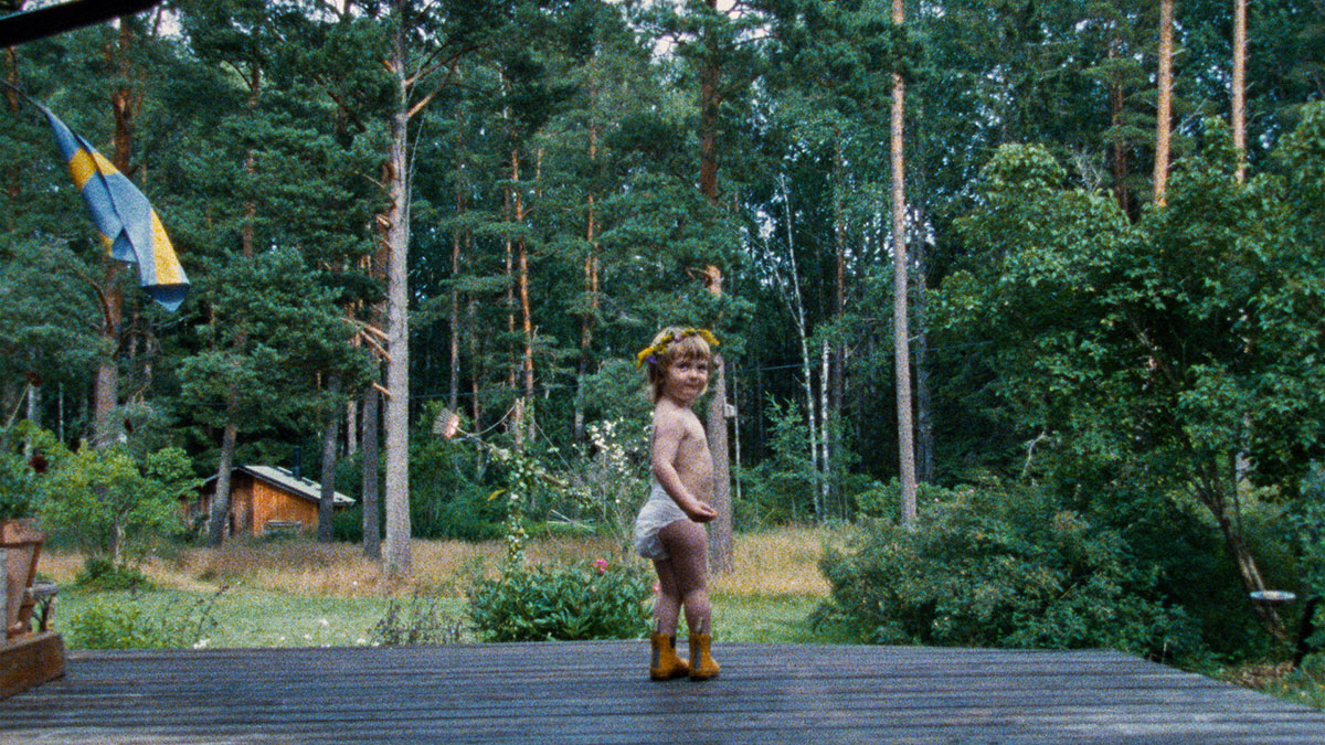 En bebis i blöja och med en blomsterkrans på huvudet står på en veranda med tallskog i bakgrunden.