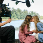 Två sittande kvinnor kyssen varandra framför en sjö. Filmkamera och mikrofon i förgrunden.