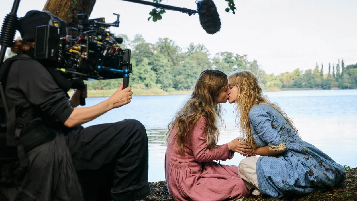 Två sittande kvinnor kyssen varandra framför en sjö. Filmkamera och mikrofon i förgrunden.