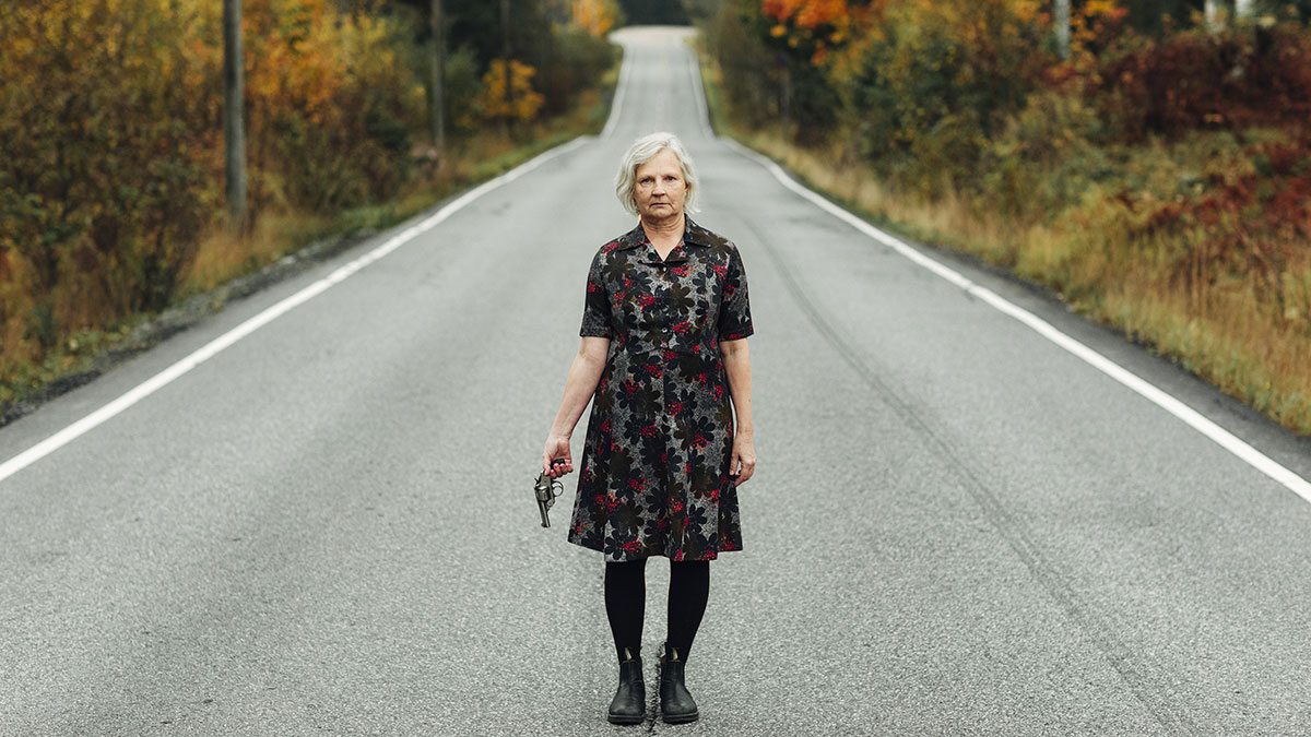 Kvinna med revolver i handen står på en bilväg med skog i bakgrunden.